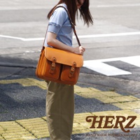 日本HERZ職人手作經典雙扣郵差包，由一位女士肩背，包款為焦糖色皮革，設計有雙前袋和金屬釦環。