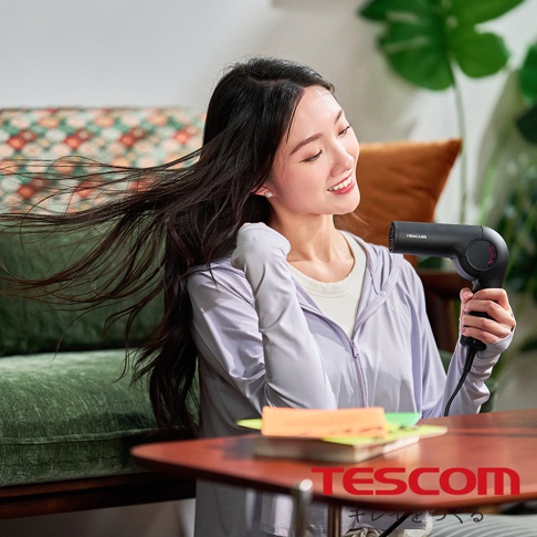 一位女性正在使用TESCOM品牌黑色摺疊式吹風機吹乾頭髮，頭髮隨風飄動，她臉上帶著愉悅的笑容。