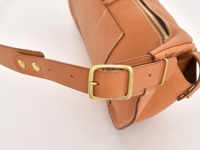 淺棕色皮革斜背包，配有黃銅色金屬帶扣和鉚釘，可調節的皮質肩帶。