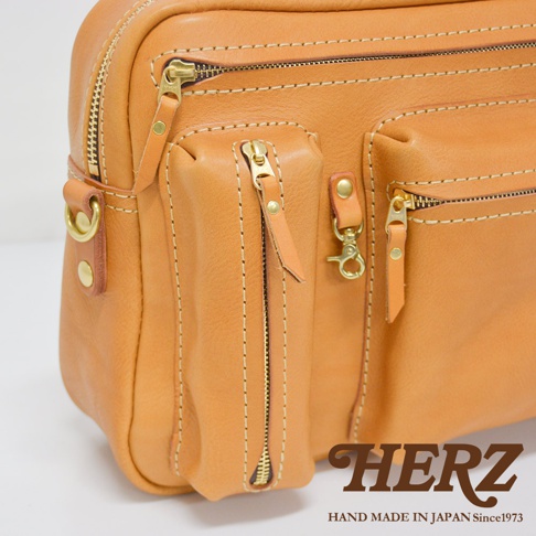 職人手作皮革兩用多功能收納單肩包，呈現自然原色皮革，搭配黃銅色拉鍊與五金配件，包身結構明顯，設有多個拉鍊口袋與皮質拉環，縫線清晰。