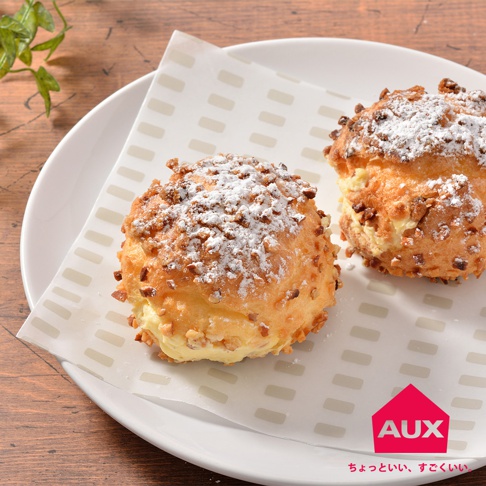 兩個奶油泡芙，上面撒著糖粉和切碎的堅果，放在印有灰色條紋圖案的白色紙上，紙放在白色盤子上，背景為木紋桌面。