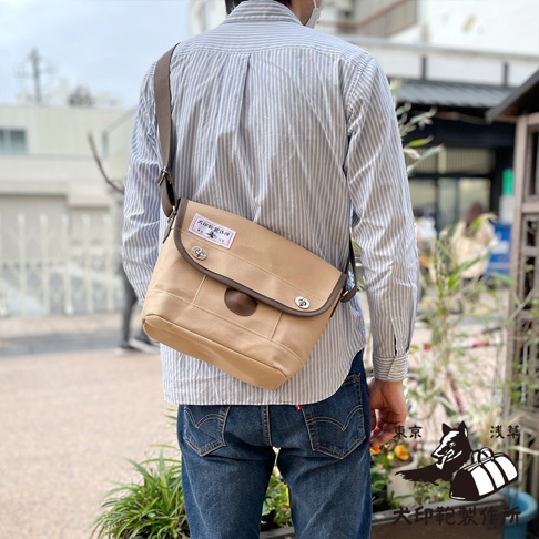 犬印純綿帆布雙扣郵差包，淺褐色帆布材質，附有深棕色皮革飾邊與標籤，可調節肩帶。