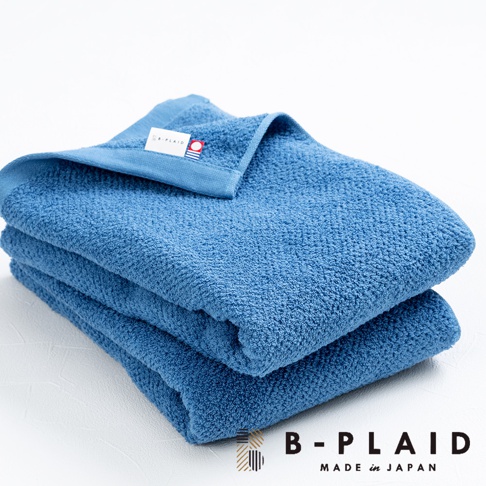 藍色毛巾堆疊，表面有細緻的鱗紋織法，左上角有品牌標籤。