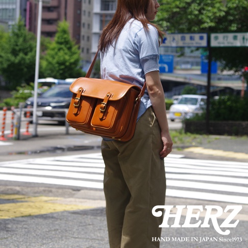 日本HERZ職人手作皮革經典雙扣郵差包，棕色皮革材質，女性斜挎於肩，呈現經典雙扣設計與精緻工藝，圖片下方有HERZ LOGO和「HAND MADE IN JAPAN Since1973」字樣。