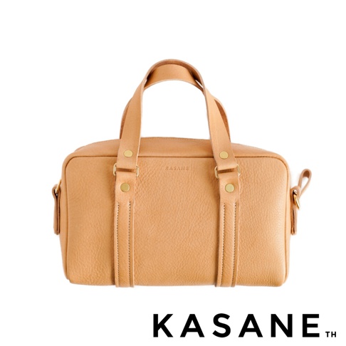 一個淺棕色的KASANE職人手作義大利皮革迷你波士頓包，正面設計有兩條垂直的皮革飾條與金色鉚釘裝飾，整體呈現簡約質感，下方印有品牌名稱。