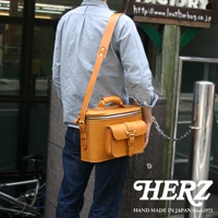模式B：一位男士身揹日本HERZ手工製作的駝色皮革相機包，包包上有兩個立體口袋，搭配銀色金屬扣環，整體呈現復古風格。