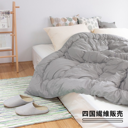 一張雙人灰色舒暖被，鋪蓋在白色床單上，被面有明顯的絎縫紋路。床頭擺放著多個靠墊，其中兩個為白色，另外兩個則有圓形圖案。床邊地上有條彩色條紋地毯，上面放著一雙灰色條紋拖鞋。