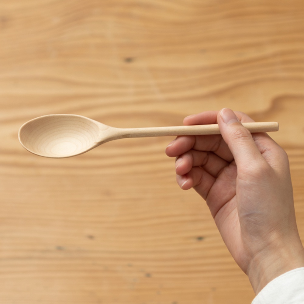 一隻手拿著一支淺色楓木湯匙，湯匙呈現自然的木紋，背景為木質桌面。