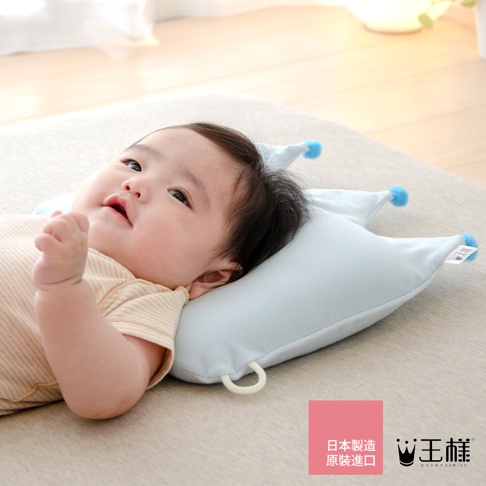 嬰兒仰躺在淺藍色皇冠造型的嬰兒枕上，枕頭上有兩個藍色絨球裝飾，枕頭一側有品牌標籤。