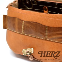 職人手作皮革鹿角扣繩郵差包，採用鞣製皮革製成，主體為淺棕色，飾有深棕色皮革拼接與縫線。包款細節處可見鹿角製成的扣環和皮革細繩。