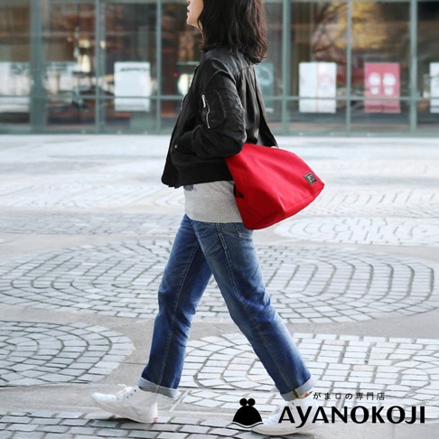 一名女子穿著黑色飛行夾克和牛仔褲，肩上揹著一個鮮紅色的 AYANOKOJI 口金包，走在鋪滿石磚的街道上。