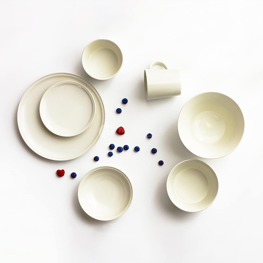 一套米白色的美濃燒餐具，包含大小盤、碗及馬克杯，散落在白色背景上，部分點綴藍莓與覆盆子。