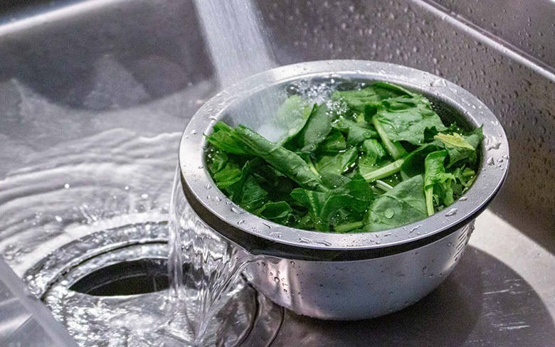 不鏽鋼濾水網盆裝滿綠葉蔬菜，放在水槽中，水正從盆中流過。