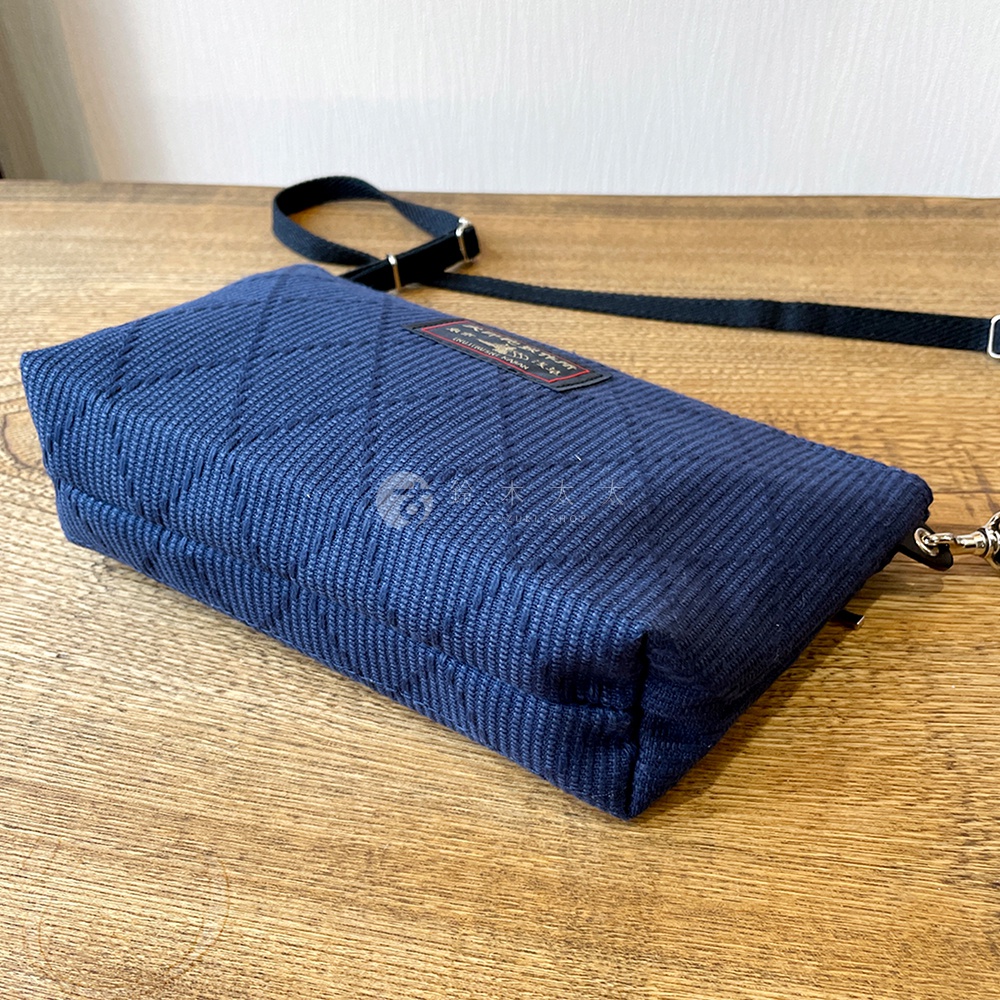 深藍色刺子織紋斜背包，帶有黑色可調式背帶，包身表面有菱格紋縫線，側面有一個品牌標籤。