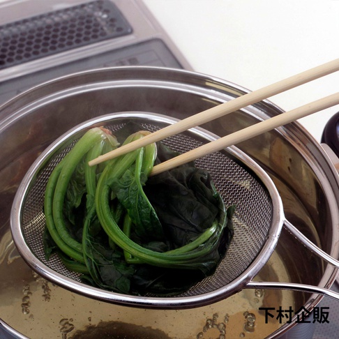 一個橢圓形的不鏽鋼濾網，網孔細密，裝有燙熟的綠色蔬菜（菠菜），由兩根木筷夾著，置於鍋中。