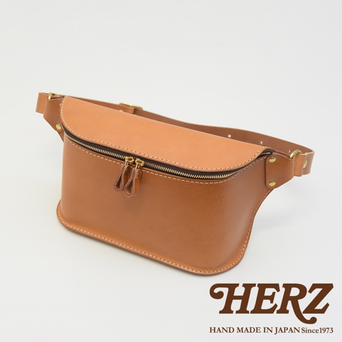 職人手作皮革立體斜背包，採用淺棕色皮革，設有雙拉鍊開口，搭配可調節式皮帶，整體設計簡約實用。