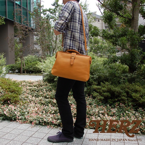 職人手作皮革軟式杜勒斯包，黃褐色皮革材質，附有金屬扣環的翻蓋設計，可斜背或肩背。