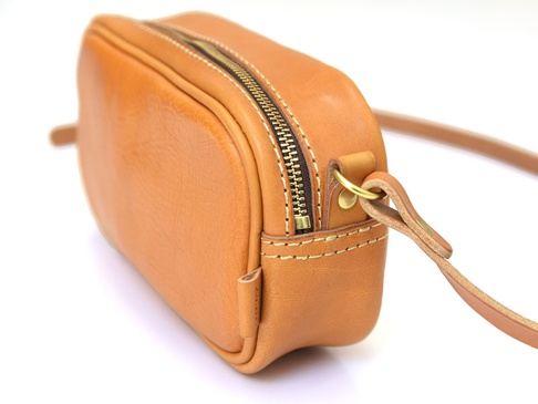淺棕色皮革製成的手工隨身單肩小包，具有金屬拉鍊和可調節的皮革背帶，透過縫線裝飾。