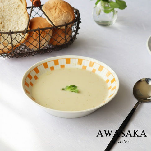 一個裝有奶油湯的白色圓形盤子，盤邊有橘色格紋裝飾，湯上點綴著綠色薄荷葉。盤子旁有一把黑色手柄的銀色湯匙。背景是白色桌布，左側有一個黑色線編織籃，籃裡有幾片吐司和麵包。