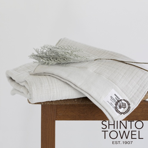 兩條淺米色雙面紗布毛巾疊放，其中一條毛巾上有一束乾燥的白色植物裝飾。毛巾表面有細緻的格紋織紋，其中一條毛巾的邊緣縫有印有品牌標誌的白色標籤。