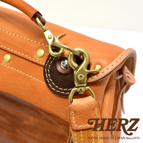 鞣製皮革製成的郵差包，帶有鹿角狀扣環和鉤扣，搭配皮革提把和肩帶，整體為淺棕色，細節處使用深棕色皮革點綴，並以縫線和金屬配件強化結構。