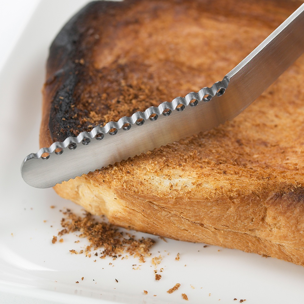 日製不鏽鋼奶油刀，刀刃鋸齒狀，帶有圓孔設計，正切在一片帶有焦黑邊緣的烤吐司上，吐司表面散落著麵包屑。