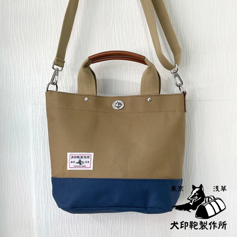 犬印純棉帆布兩用雙色托特包，主體為卡其色，底部為海軍藍。包款配有兩條可拆卸肩帶，一條是帆布材質，另一條是皮革提把。包面中央有品牌標籤。