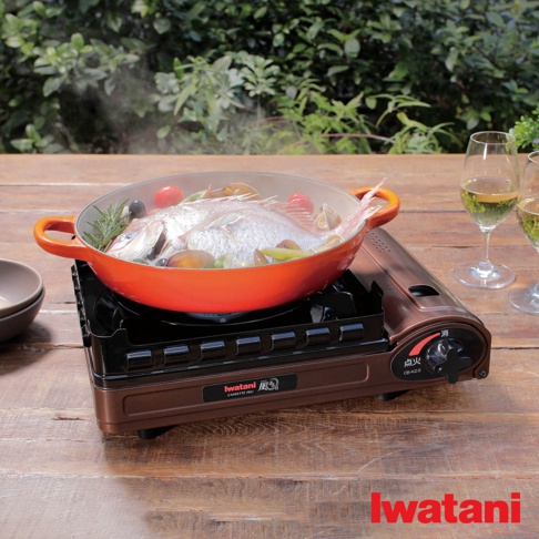 一台棕色金屬材質的雙重防風卡式爐，爐上放置著橘色鑄鐵鍋，鍋內有整條魚、蛤蜊、小番茄、蔥段等食材正在烹煮，鍋邊有蒸氣冒出。