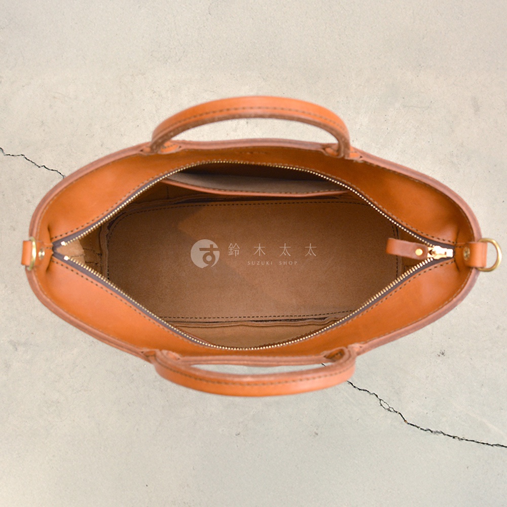 一個棕色皮革托特包，從上方俯視，包包開著拉鍊，裡面可以看到 SUZUKI SHOP 的品牌標誌。