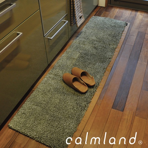 一張綠色、長條形的廚房地墊，上面擺放著一雙棕色皮革拖鞋。地墊材質蓬鬆，鋪設在木質地板上，旁邊是廚櫃。