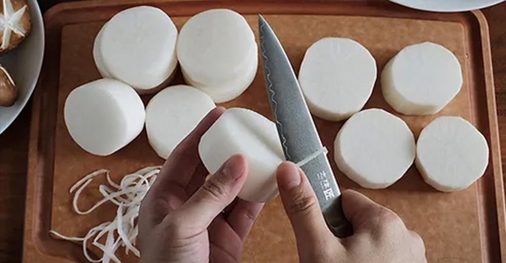 一把帶有波浪紋刀刃的銀色主廚刀，正在處理切成圓片的白蘿蔔。刀柄上有中文文字。