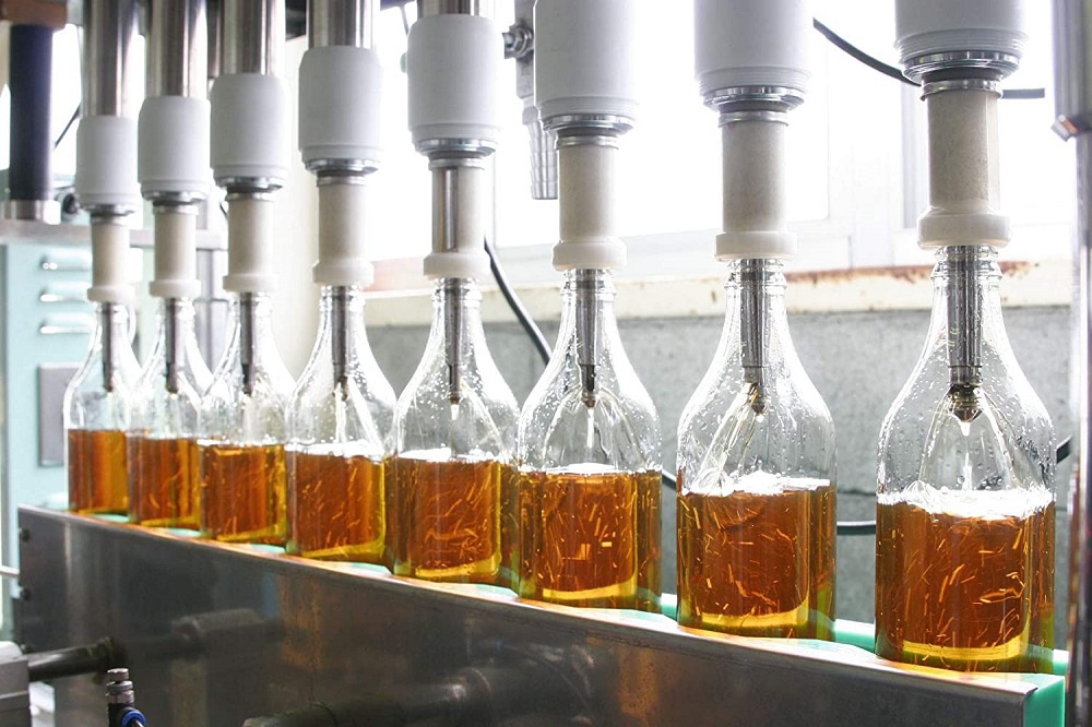 多個透明玻璃瓶盛裝著琥珀色液體，瓶口處有銀色金屬填充管，瓶身有水珠，產品正在自動化生產線上進行填充作業。