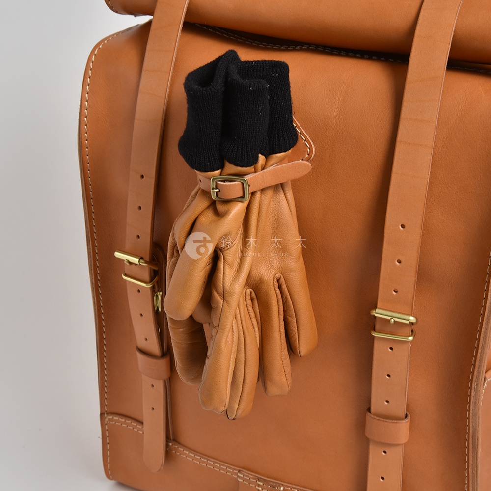 職人手作皮革後背包，展示棕色皮革材質，附有黑色針織手套，皮革手套以小皮帶和金屬扣固定於背包肩帶上。