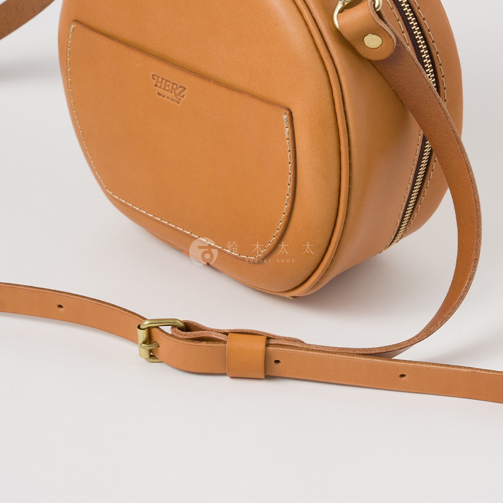 日本HERZ品牌手工製作的淺棕色皮革圓形斜背包，特寫其前方口袋及可調節的皮革背帶與金屬扣環。