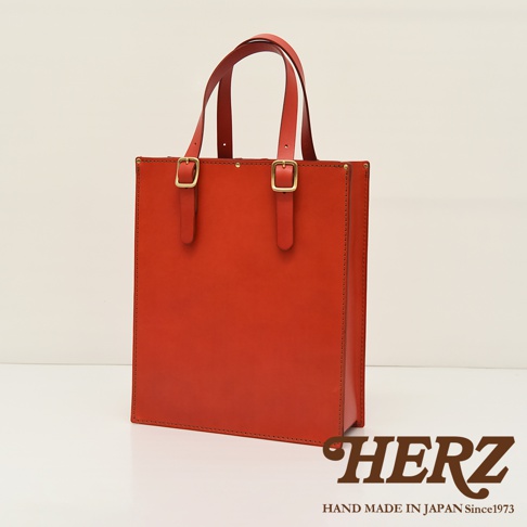 橘紅色皮革製成的直立式托特包，配有兩個可調節的皮質提把，提把上有金色調節扣，包身側面有縫線裝飾，整體呈現簡約風格。