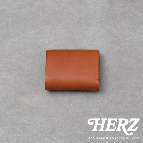 日本HERZ職人手作皮革迷你短夾，選用棕色Minerva Box皮革製成，正面呈現簡約平滑的質感，右側可見精緻的車縫細節，整體採日本手工製作，右下方印有品牌標誌及產地資訊。