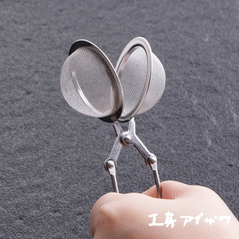 銀色不鏽鋼製的圓形泡茶器，網狀濾網，附有可開合的夾子設計，由手持。