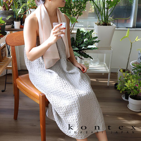 一位女性坐姿，身穿淺灰色棉麻混紡平口鬆緊浴巾浴裙，身上搭著一條淡粉色毛巾，手持一杯水，背景為室內綠植。