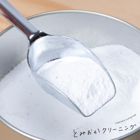 銀色金屬勺子盛滿白色細緻的洗衣粉，部分洗衣粉散落在銀色金屬罐中，罐內洗衣粉可見細小的藍色顆粒。