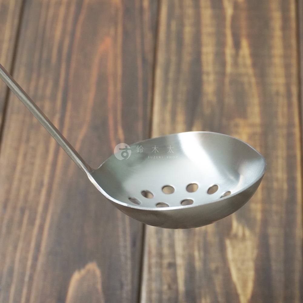 銀色不鏽鋼萬用瀝水湯勺，勺面有圓形瀝水孔，勺柄細長，置於深褐色木紋背景上。