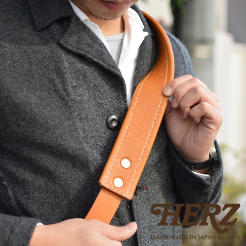 職人手作淺棕色皮革復古翻蓋郵差包，搭配白色縫線及銀色金屬釦，由穿著深灰色外套的男性肩背。