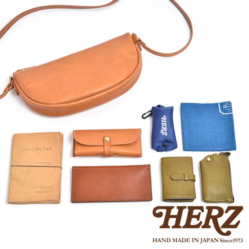 日本HERZ品牌半圓形皮革馬鞍包，搭配多款品牌周邊小物，包含錢包、記事本、鑰匙包、收納袋及藍色印花方巾，展現職人手工製作的日式風格。