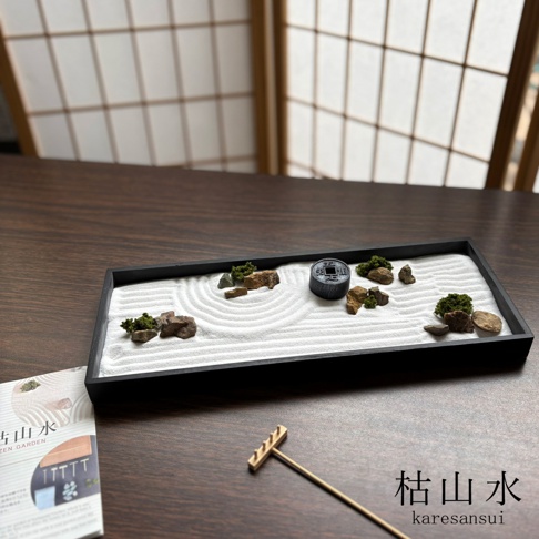 一張桌面上擺放著日本枯山水模型，黑色長方形托盤內鋪滿白色細沙，沙面劃出環形紋路，裝飾有岩石、微縮青苔植栽以及一個刻有文字的圓形飾物，旁邊放著一支小型木製耙子，左下角有一本說明書，右下角則標示著枯山水及英文書寫的karesansui字樣。