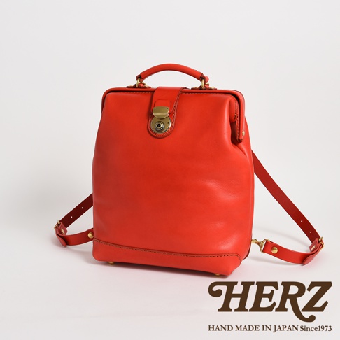 這是一款由日本品牌HERZ職人手工製作的亮紅色皮革後背包，包款採醫生包式的開口設計，正面配有黃銅材質的圓形轉扣，頂部設有堅固的皮革提把，並附有可調節的雙肩背帶，包底裝有四顆金屬腳釘，整體呈現簡約復古的質感。