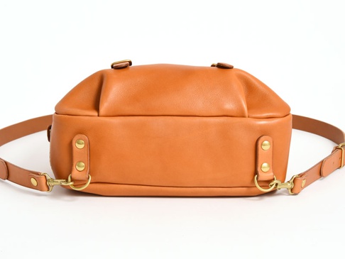 經典雙扣後背包，採用棕褐色皮革製成，設有兩條可調節的皮革背帶，以黃銅色雙扣和鉚釘固定，設計簡約。