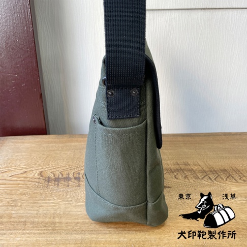 軍綠色純棉帆布雙扣郵差包，側邊視角，包身有外袋，黑色織帶肩帶。