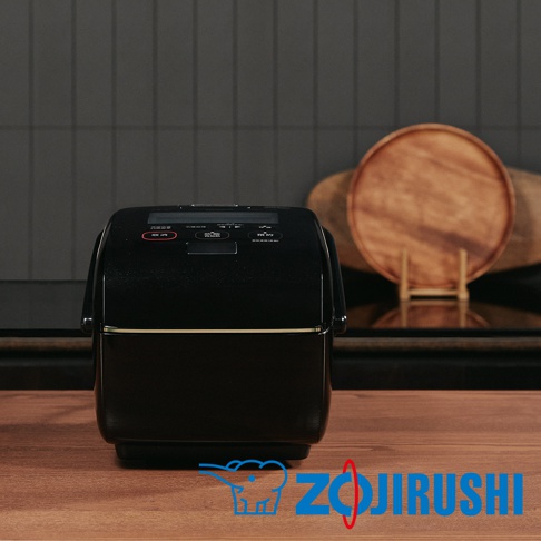 黑色炎舞壓力IH電子鍋，具備數位顯示螢幕與多個功能按鈕，頂部有金色飾條。置於木紋桌面上，背景為灰色條紋牆面與兩個木製圓盤。