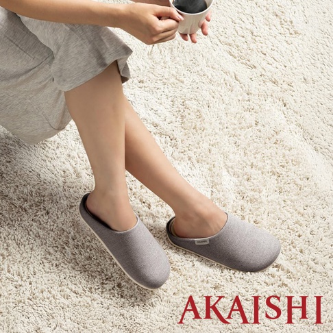 一雙灰色布面包覆式室內拖鞋，鞋底為淺色，鞋舌上有品牌標籤，展示於白色長絨毛地毯上。