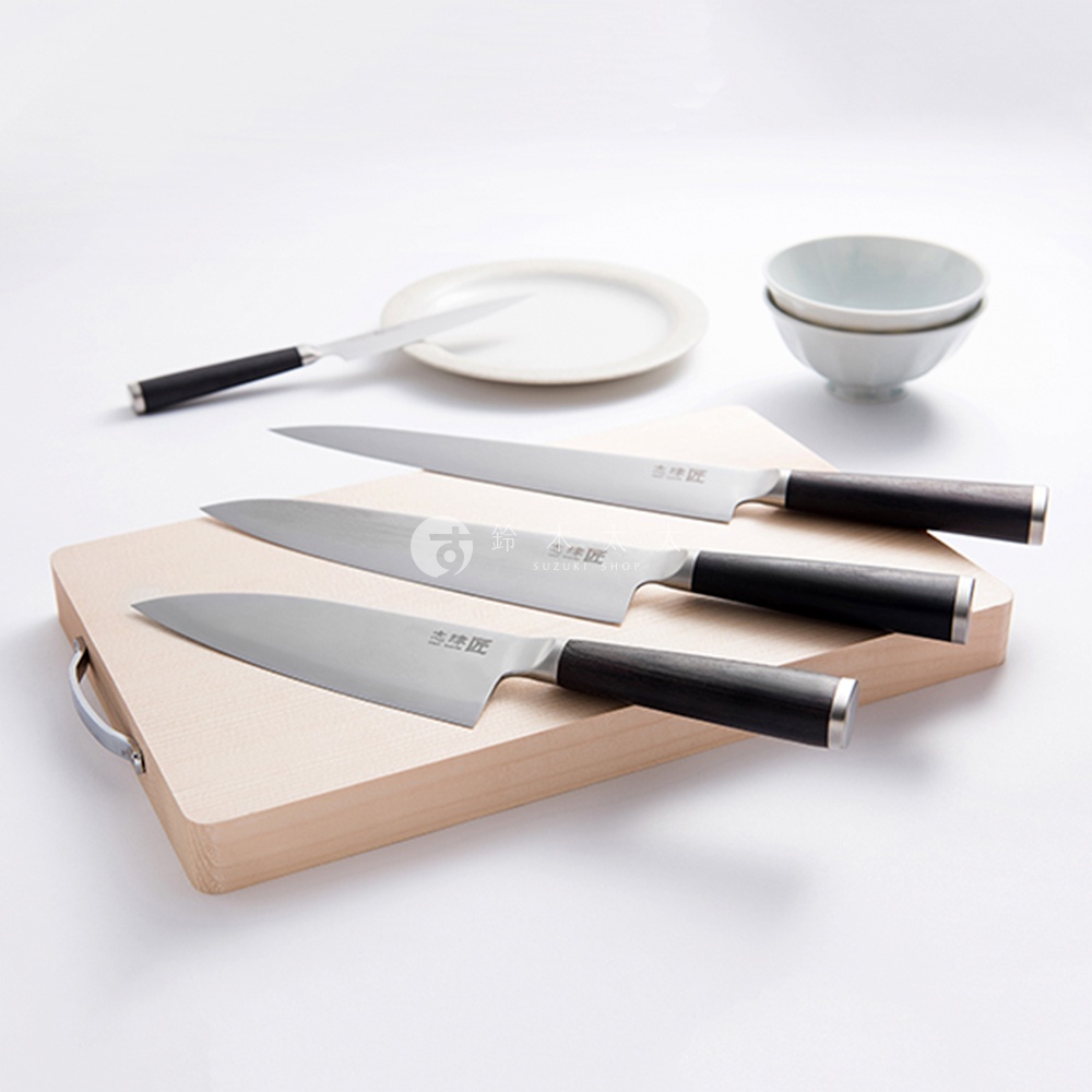 三把銀色金屬刀刃、黑色握柄的廚房刀具，其中兩把置於木質砧板上，另一把斜放於背景的白色盤子旁，另有兩個白色小碗。