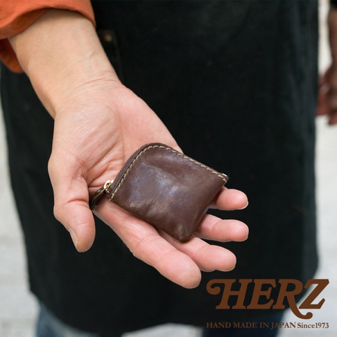 這是一張日本HERZ品牌職人手作皮革零錢包的展示圖，畫面呈現一隻手掌平托著一個深棕色的小巧正方形皮革收納袋，拉鍊位於邊角處，皮革表面具有自然的紋理與色澤，圖片右下角標註有HERZ品牌名稱以及HAND MADE IN JAPAN字樣。